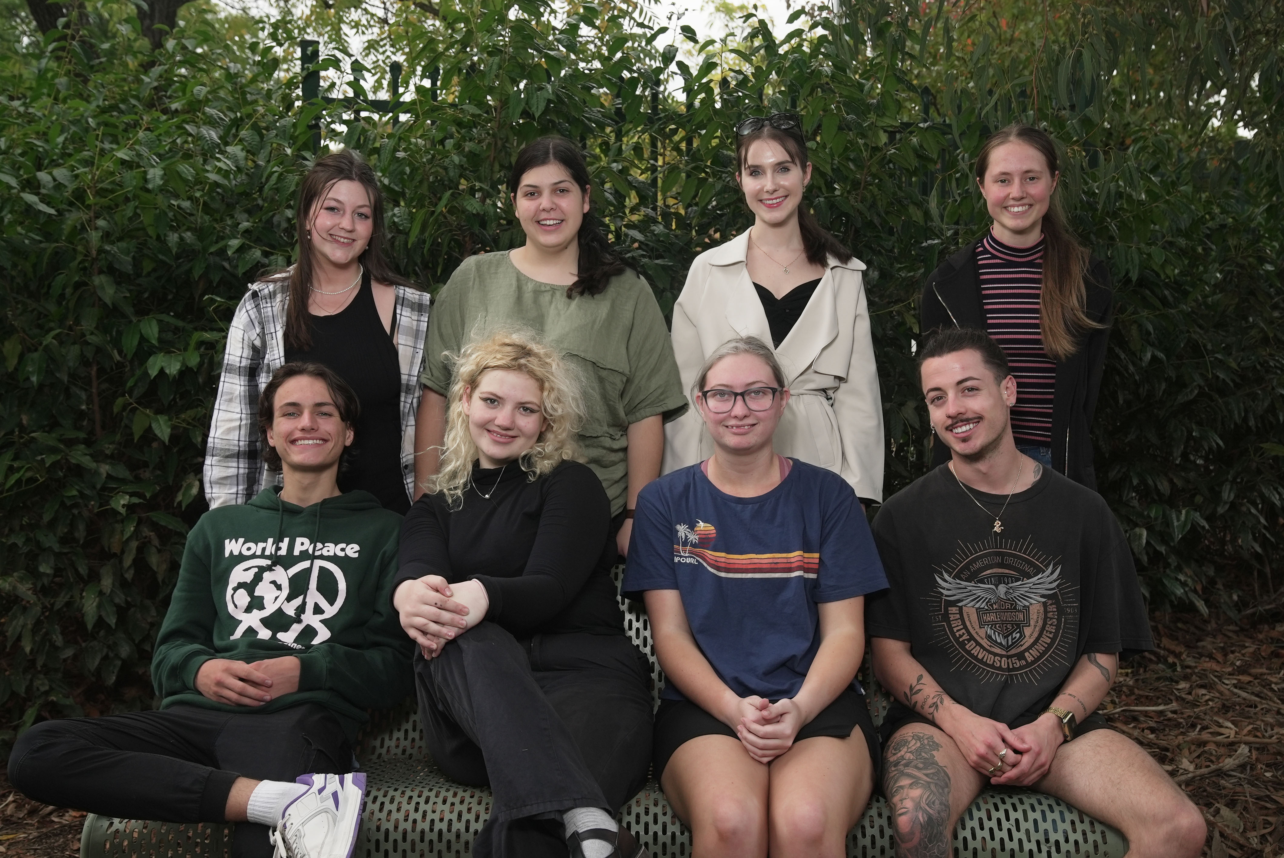 Eight people, four standing up and four sitting down on bench smiling together