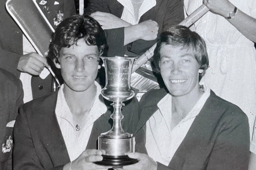 Two Australian women's cricketers hold an ODI World Cup trophy, as their teammates stand around them.