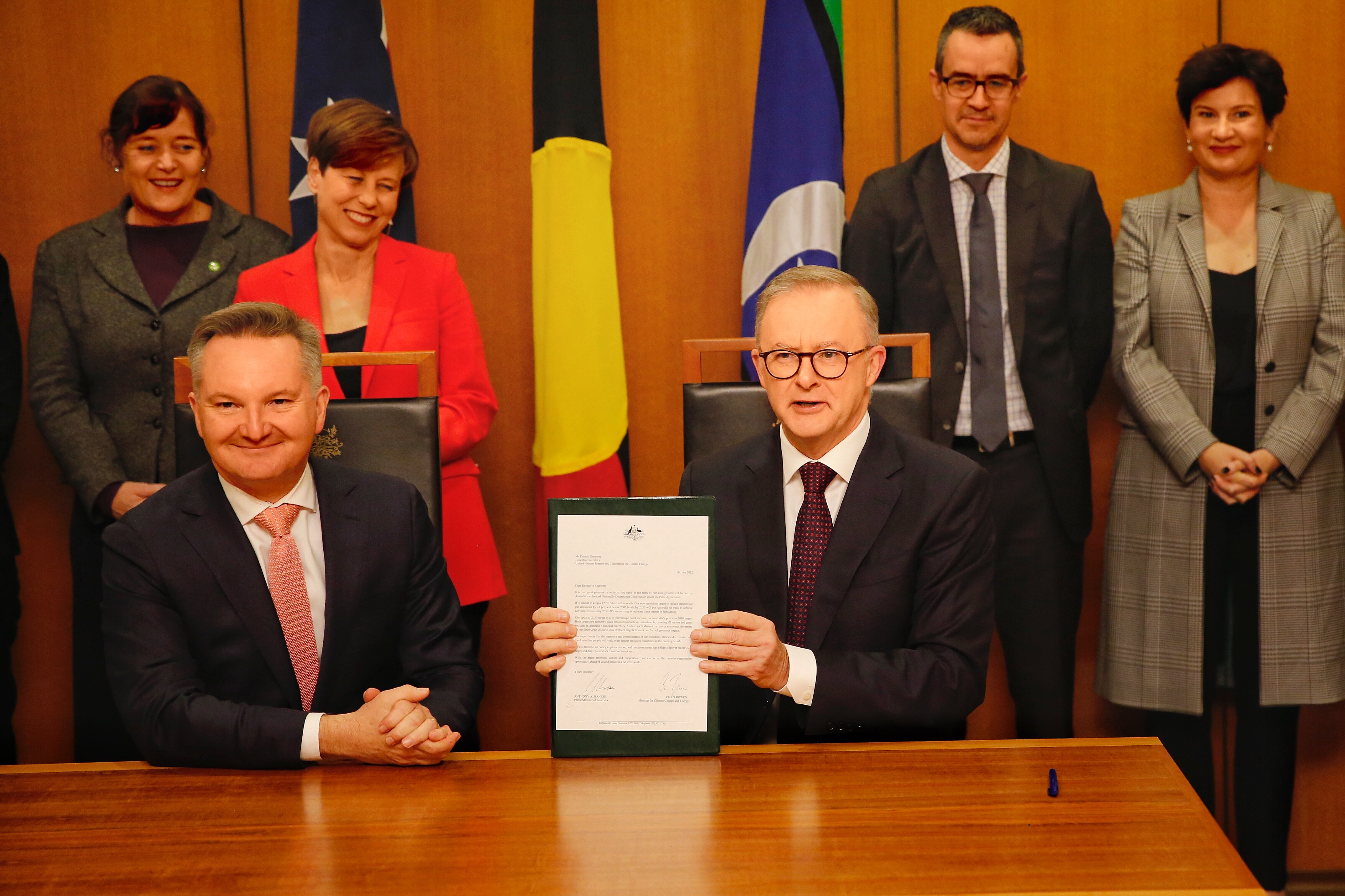 Albanese holds up a framed piece of paper, sitting beside a smiling Chris Bowen, with a coterie of people behind him.
