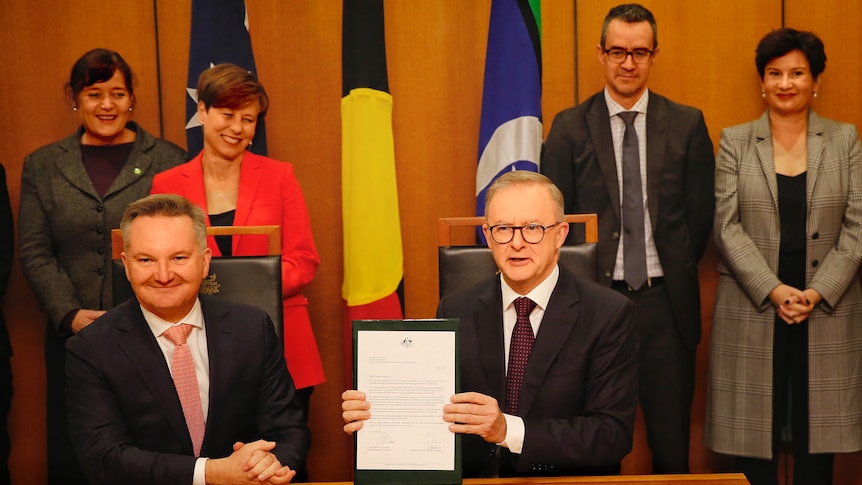 Albanese holds up a framed piece of paper, sitting beside a smiling Chris Bowen, with a coterie of people behind him.