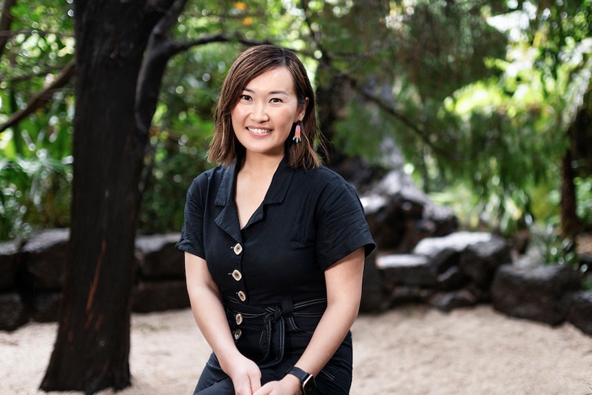 Tammy Huynh smiles in a black dress with a lush greenery in the background. 