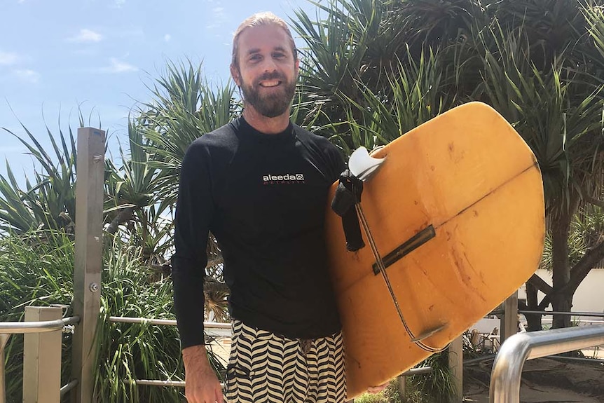 Surfer Luka Carstens-Hey stands on the sand in his rashie and boardshorts with his surfboard on the walkway to Burleigh Beach.