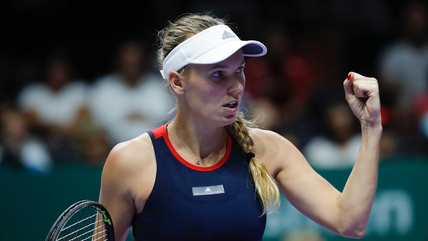 A female tennis player wearing a blue dress with red trim and a white visor cap clenches her fist