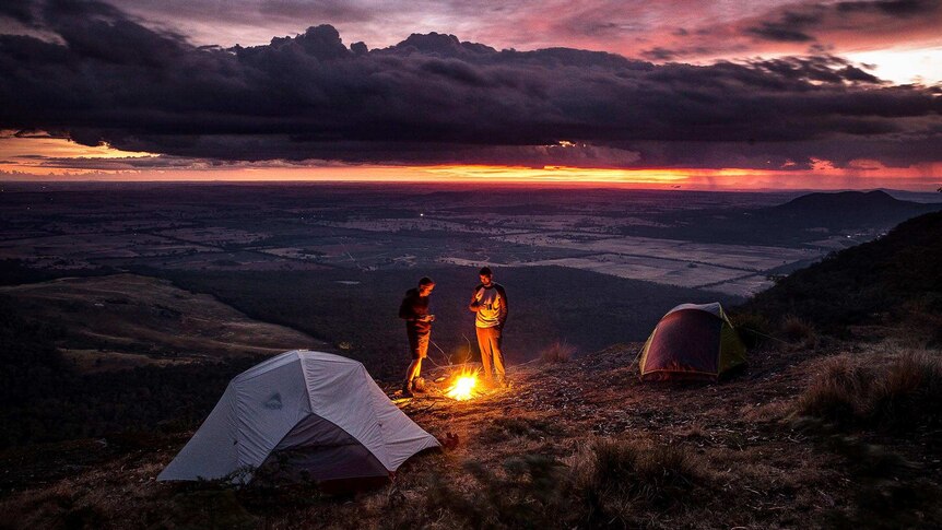 Two men stand next to a campfire and their tents on the summit of a hill at dusk, with a storm moving across the landscape below