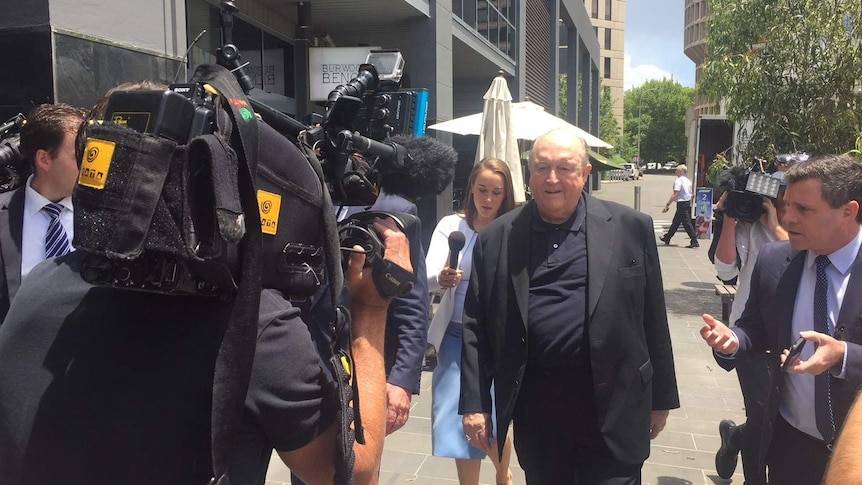 An elderly man in a black jacket walking into court flanked by journalists and news cameras.