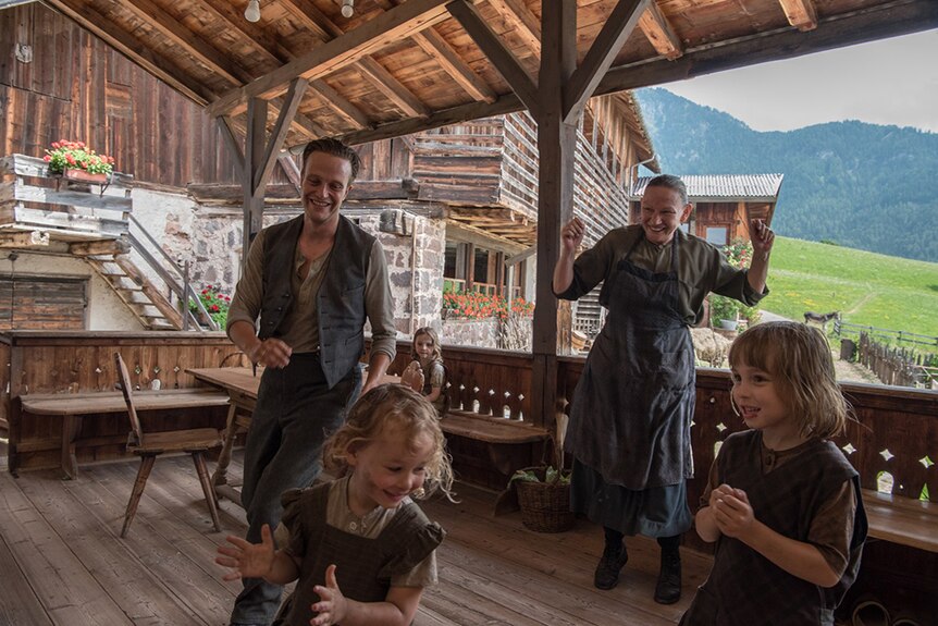 A man and his mother in peasant clothing smile and watch on as young daughters play on wooden deck in Austrian countryside.