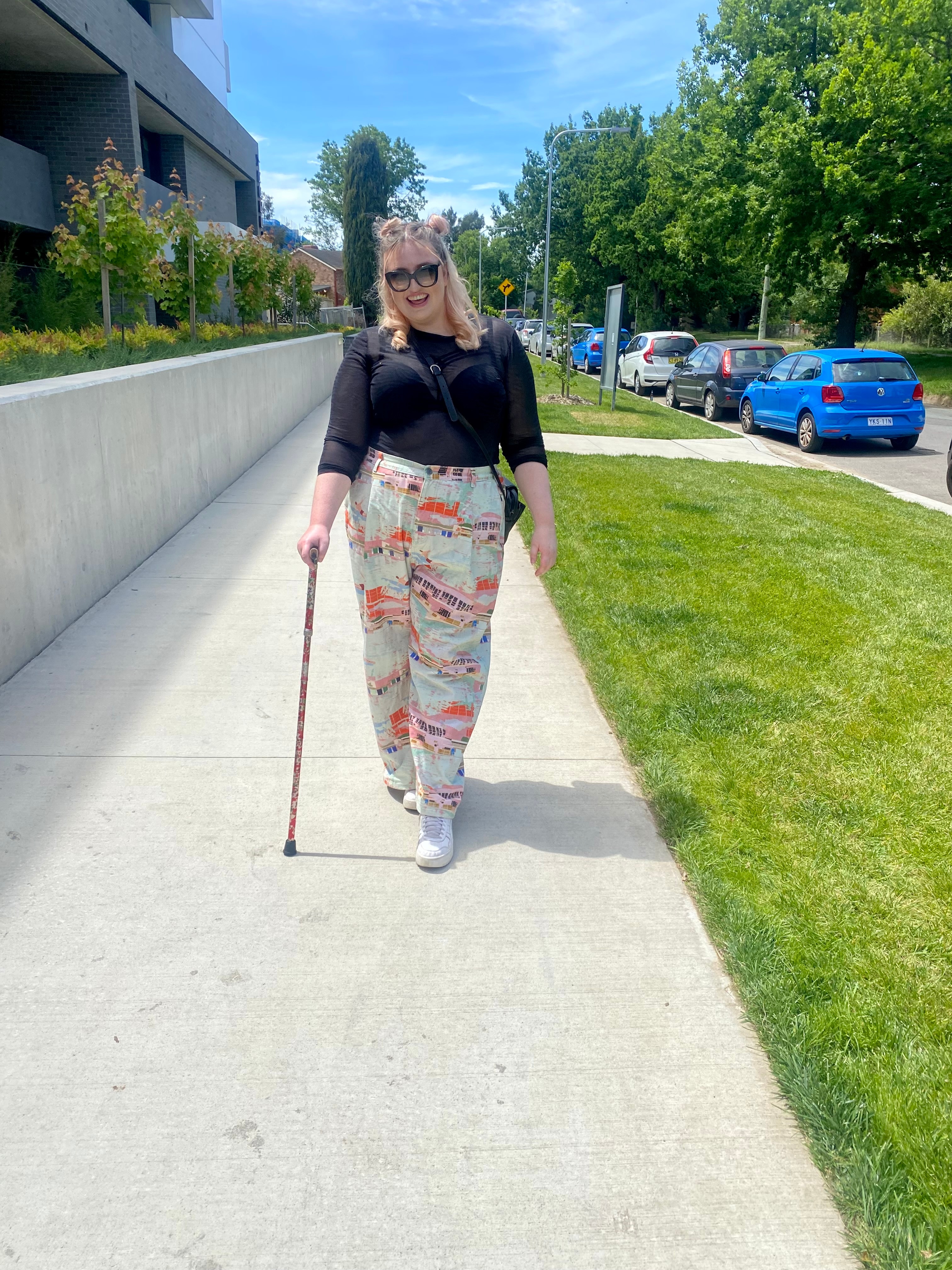Jovem funky sorrindo para a câmera. Ela está segurando uma bengala. Ela tem calças estampadas e laços espaciais no cabelo. 