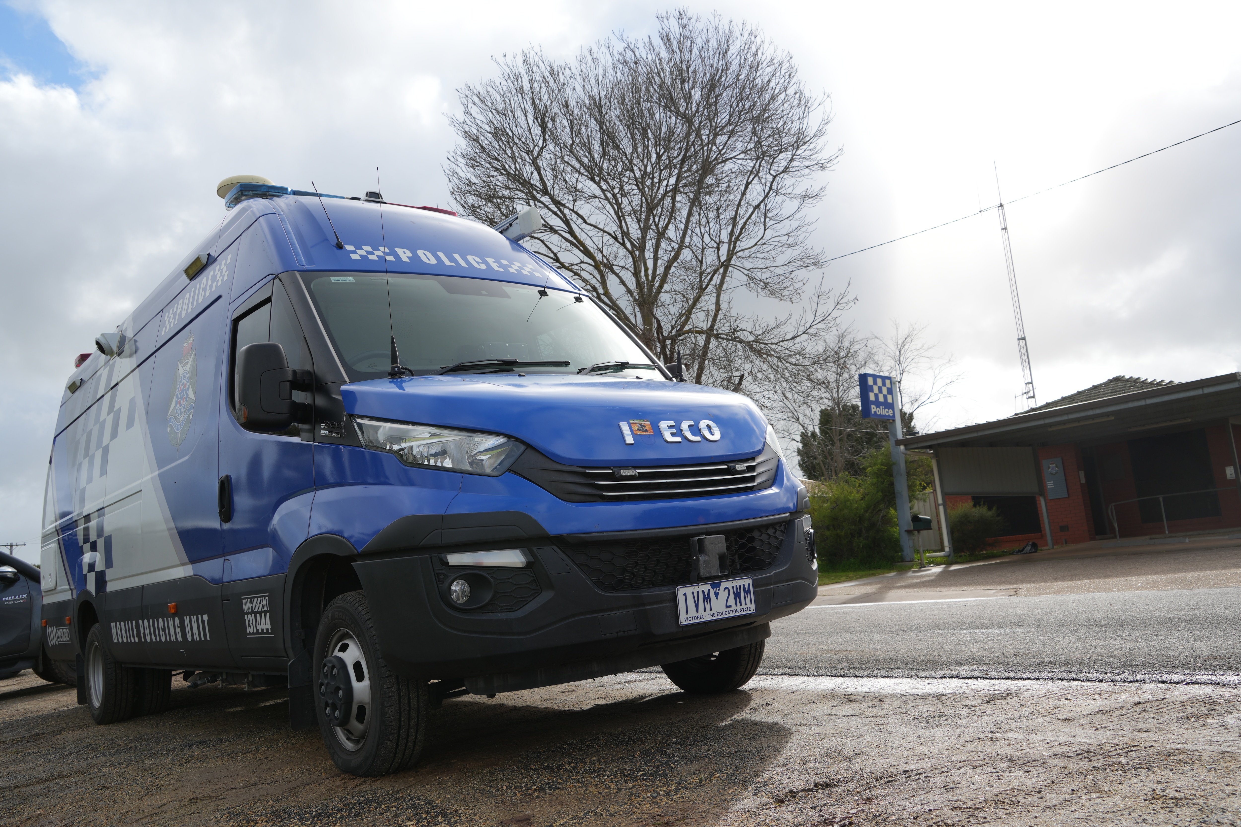 The front of a blue police van on the road. A bare tree is in the background.