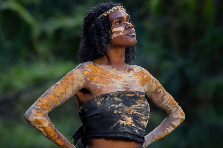 Darumbal and Torres Strait Islander woman, Kayleen Adidi, stands proudly with her hands on her hips in traditional yellow paint.