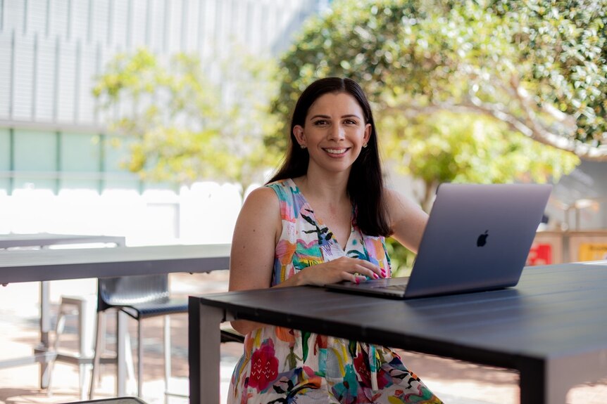 a woman sits at an outdoor table smiling with an open laptop in front of her