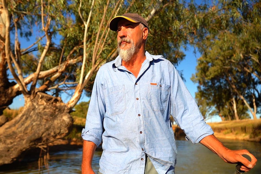 A Caucasian man with grey beard, wears a cap, blue shirt, stands near lake with trees in background, blue sky.