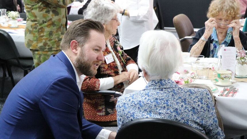A young man in a suit is sitting and speaking with three elderly women having tea.