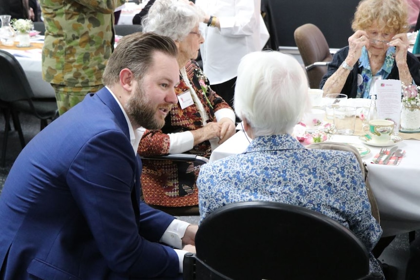 A young man in a suit is sitting and speaking with three elderly women having tea.
