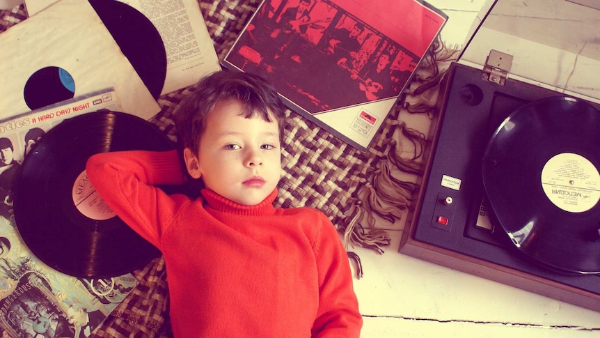 A young boy in a red turtleneck sweater lies on brown rug with Beatles vinyl records splayed next to a open record player.
