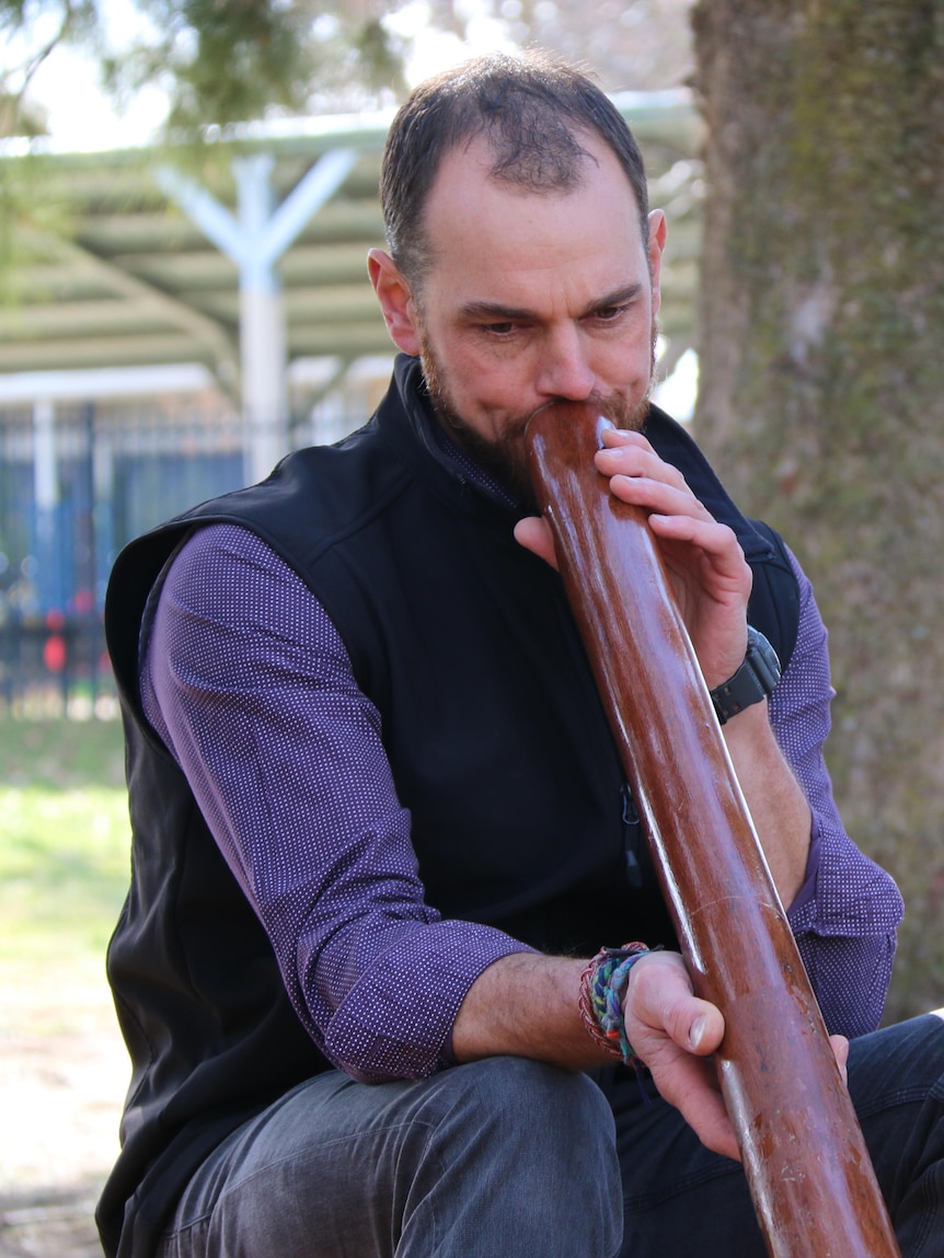 A man plays a digeridoo, outside, sitting next to school students. 