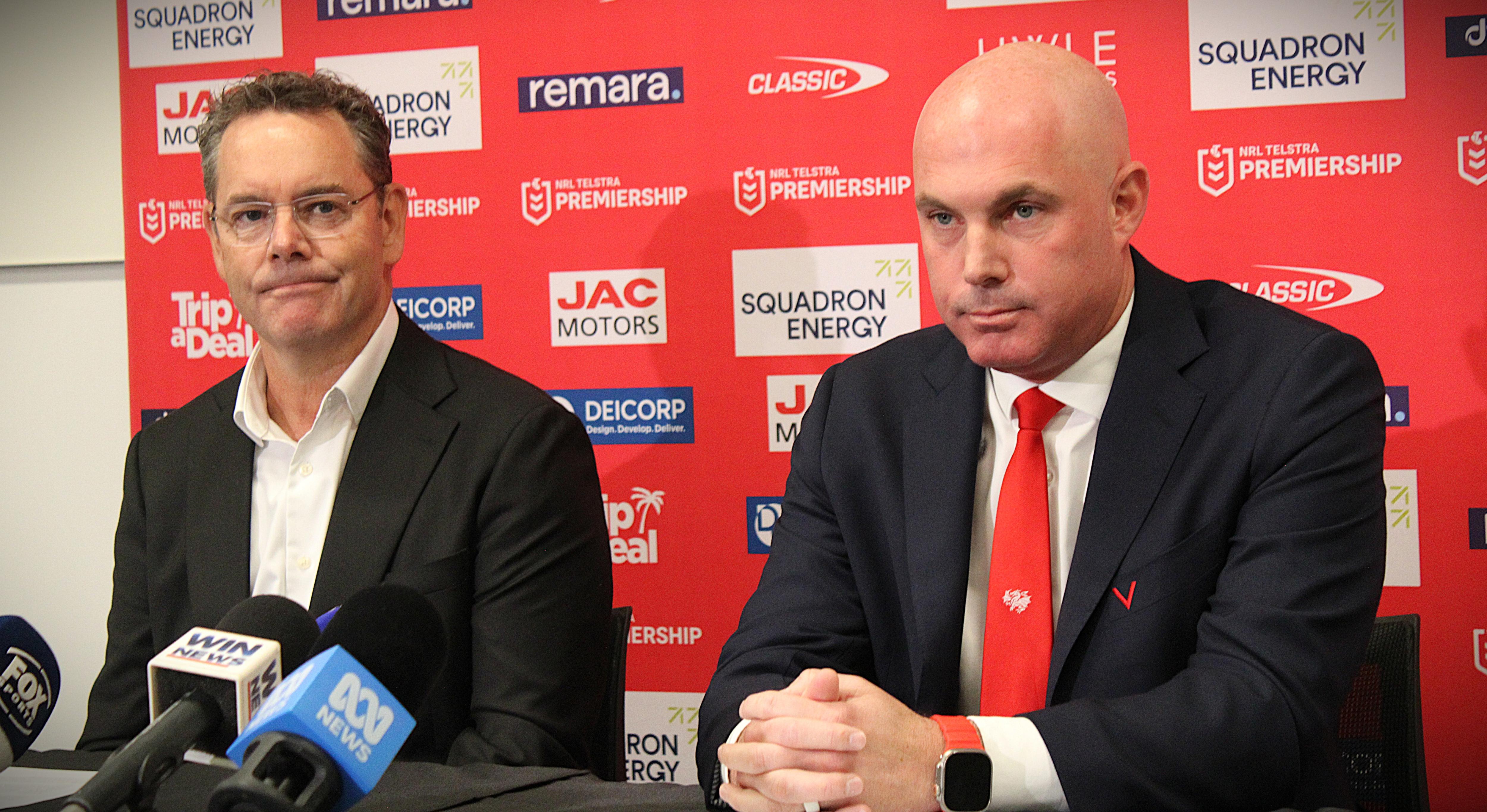 Two men sit at a table during a press conference.