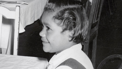 A black and white image of Lorraine Peeters sitting on a chair at a table as a young girl