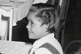 A black and white image of Lorraine Peeters sitting on a chair at a table as a young girl