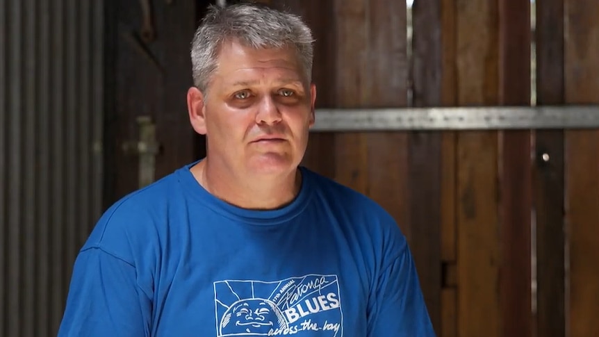 Portrait of a man in his fifties who has grey hair and a blue shirt on in a barn.