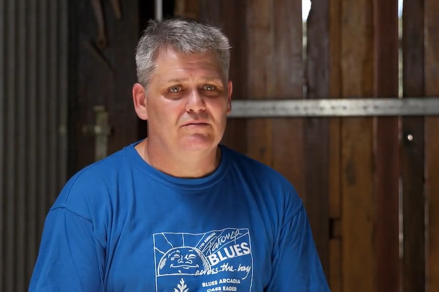 Portrait of a man in his fifties who has grey hair and a blue shirt on in a barn.