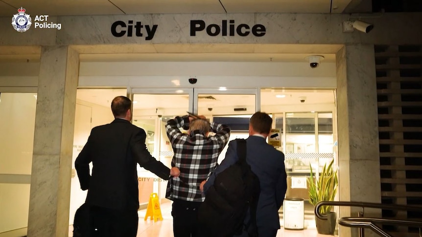 A man holding his hands above his head being led into the City Police Station.