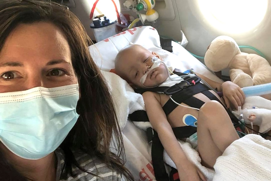 A woman wears a facemask on a plane with a sick child in an oxygen mask in a medical bed behind her.