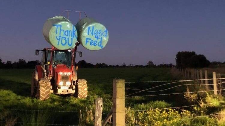 A tractor holding two hay bales with 'thanks need for feed' written on them stands in a paddock.