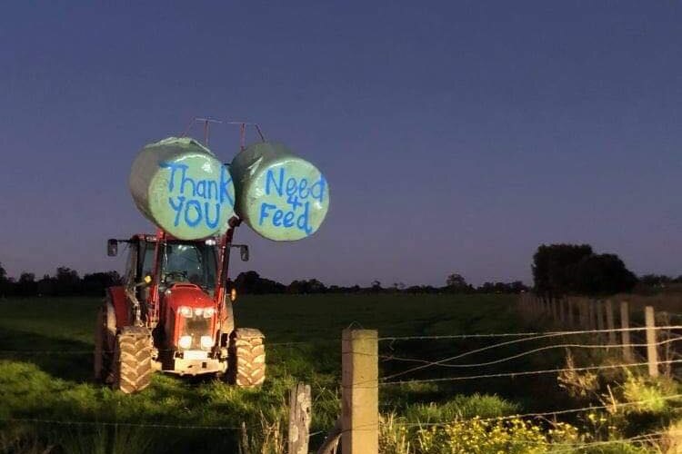 A tractor holding two hay bales with 'thanks need for feed' written on them stands in a paddock.