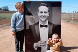 Two small children sit next to a large black and white photo of a man wearing a tuxedo suit. 