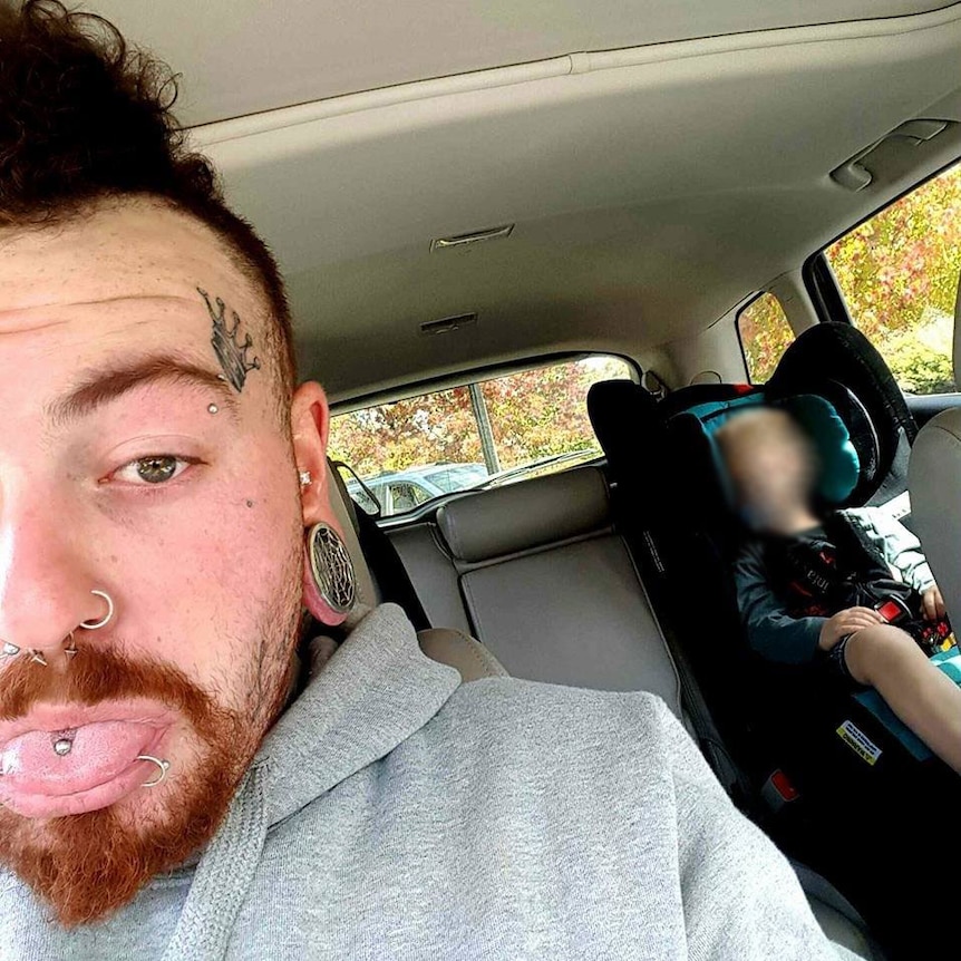 A tattooed man takes a selfie in a car. A child is asleep in the backseat.