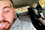 A tattooed man takes a selfie in a car. A child is asleep in the backseat.