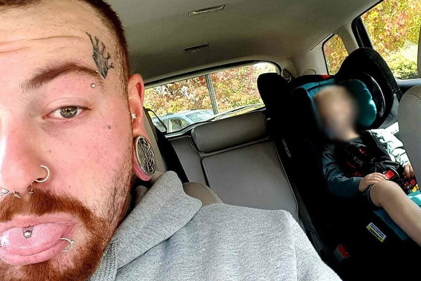 A tattooed man takes a selfie in a car. A child is asleep in the backseat.