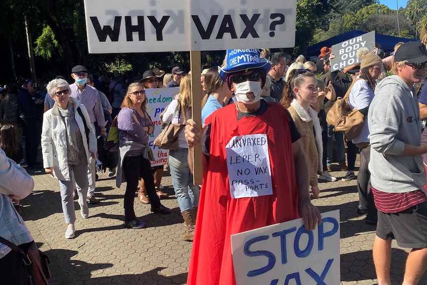 Man in mask and wearing a red cape holds a sign saying 'stop vax blackmail'.