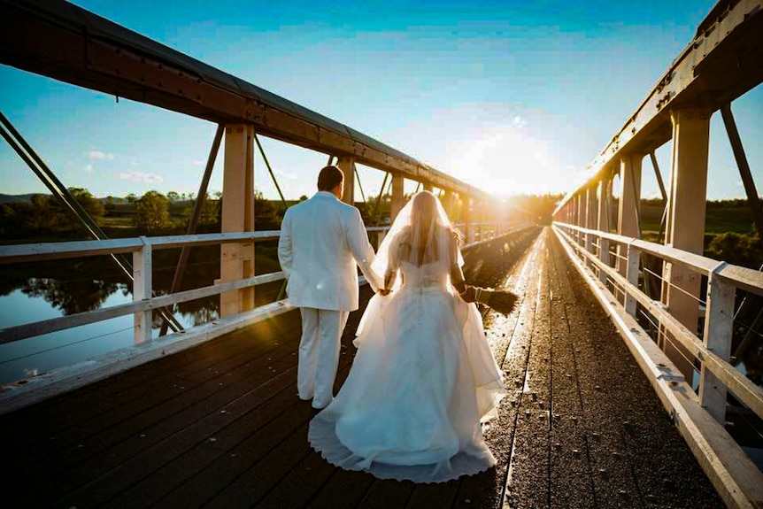 A man and woman in wedding clothes stand on a wooden truss bridge in the late afternoon golden light.