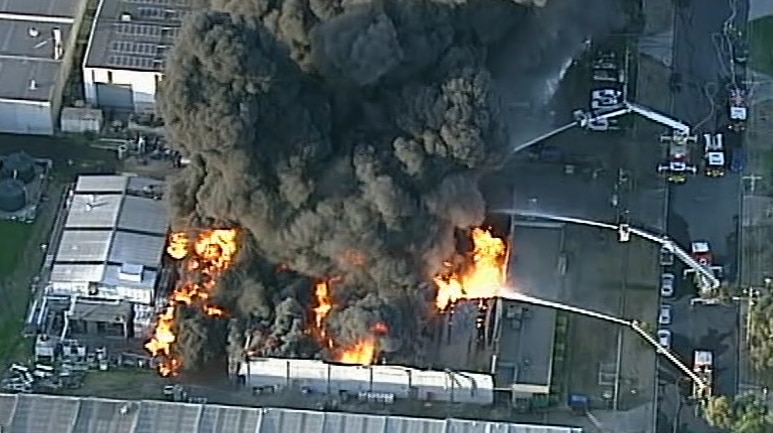Three trucks stream water over an industrial fire producing thick, black smoke.