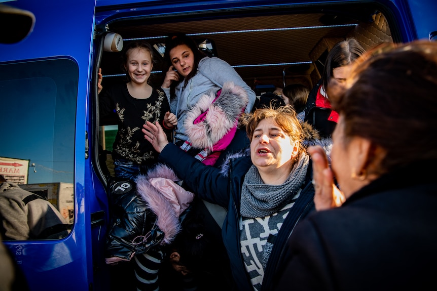 Two young children in big coats stand in a van, woman gesture outside it