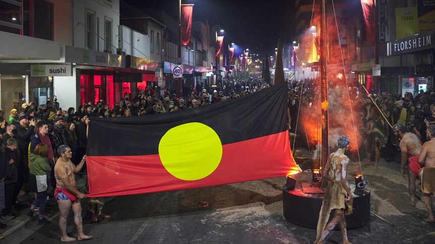 At night in a Hobart street, a young Aboriginal man holds up a Aboriginal flag, a large crowd of people gather behind the flag