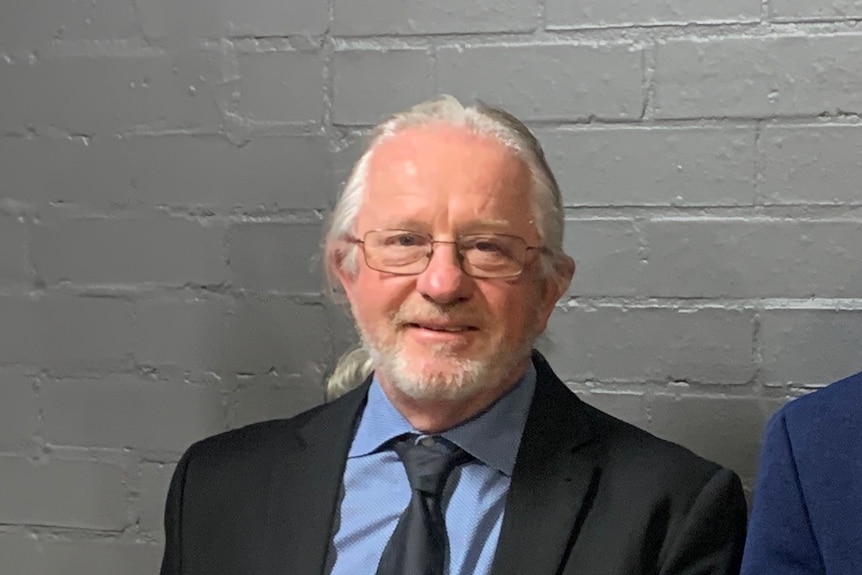 A grey-haired man in a suit standing against a grey brick wall.