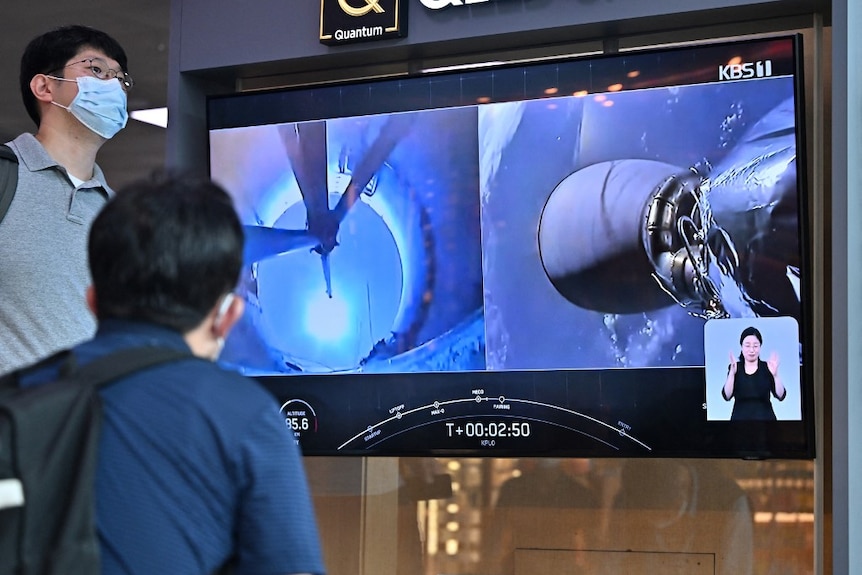 people in south korea watch and stand near a tv showing the lunar orbiter being launched into space