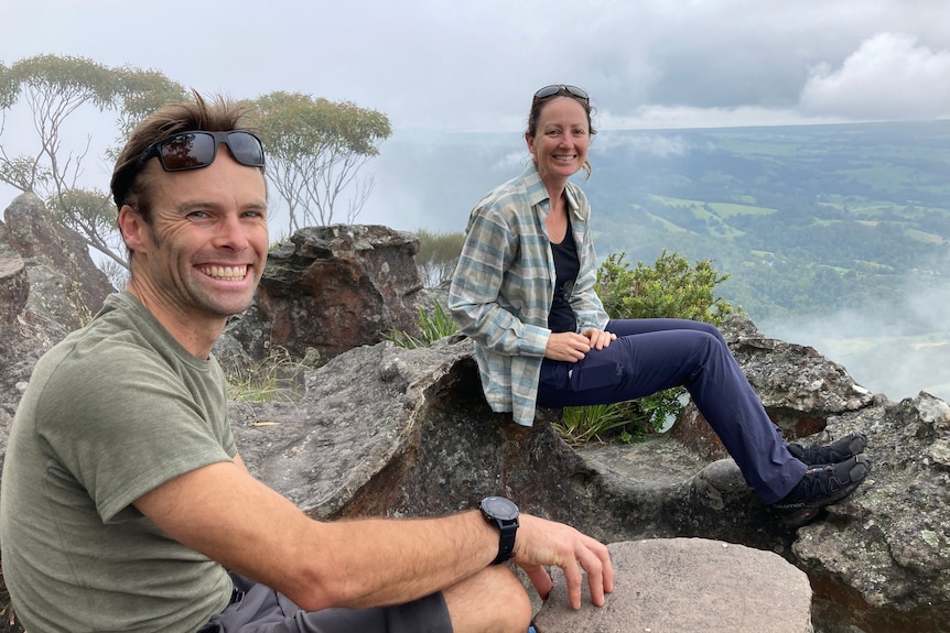 Matthew Farrell and Karen Waller smile sitting on top of rocks on the top of a mountain, with clouds below them.