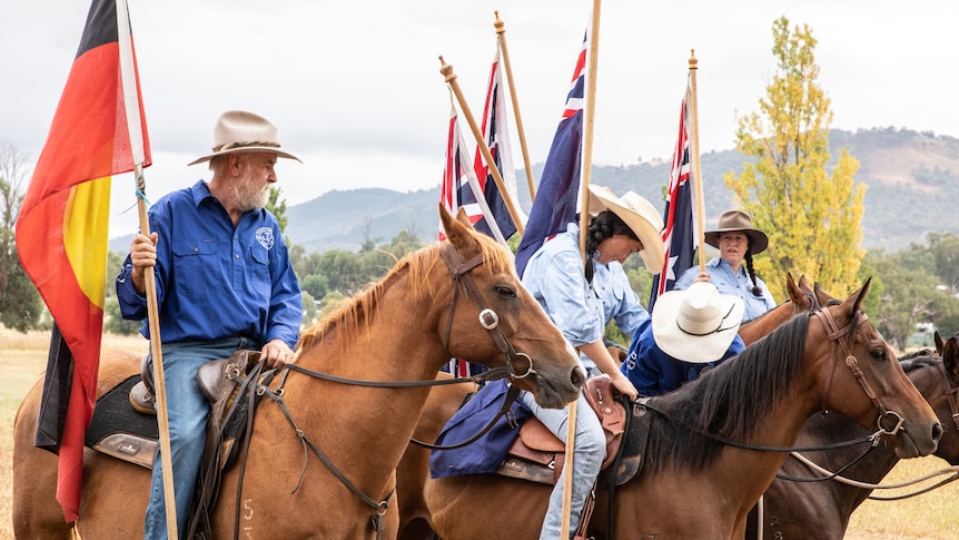 Mountain cattleman Charlie Lovick riding a horse holding the Aboriginal next to three other people riding horses with flags.