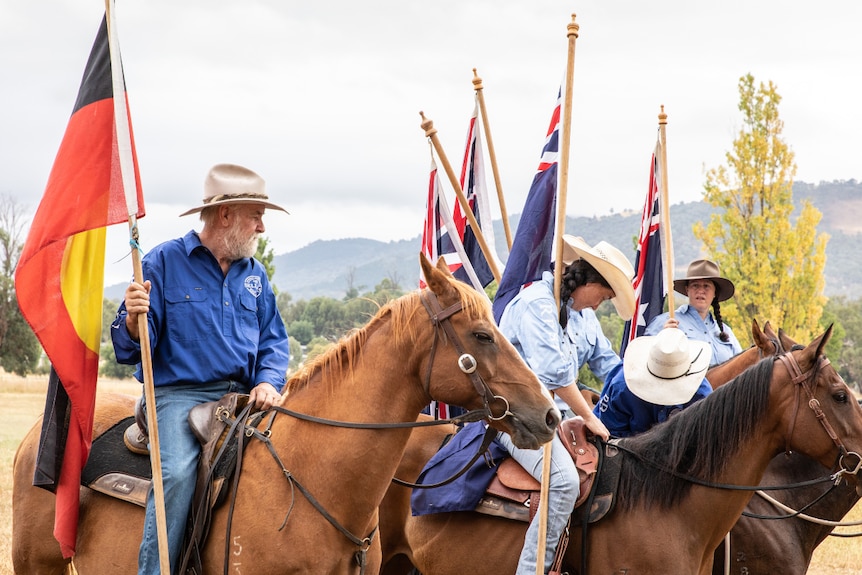 Mountain cattleman Charlie Lovick riding a horse holding the Aboriginal next to three other people riding horses with flags.
