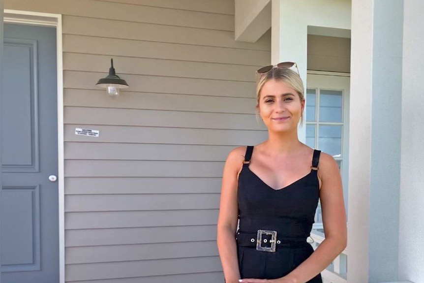 Young woman with blonde hair and black dress, standing on front patio of  house.