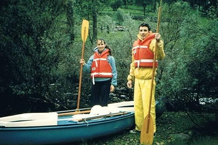 An old photo of a young man and woman in bright orange life jackets, holding oars beside a canoe. Greenery behind.