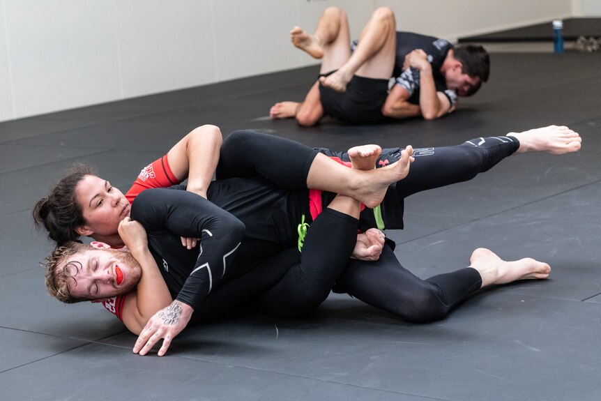 A man and woman grapple on a mat in a gym.