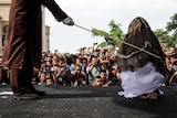 A woman is seen from behind being struck by a cane on a stage as a large crowd watches on.