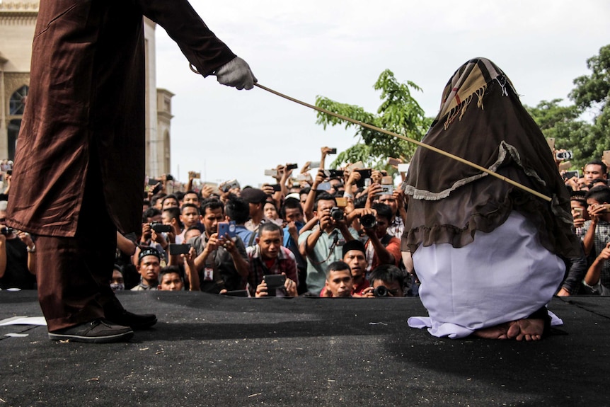 A woman is seen from behind being struck by a cane on a stage as a large crowd watches on.