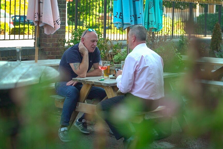 Two men sit at a wooden table at a pub beer garden.