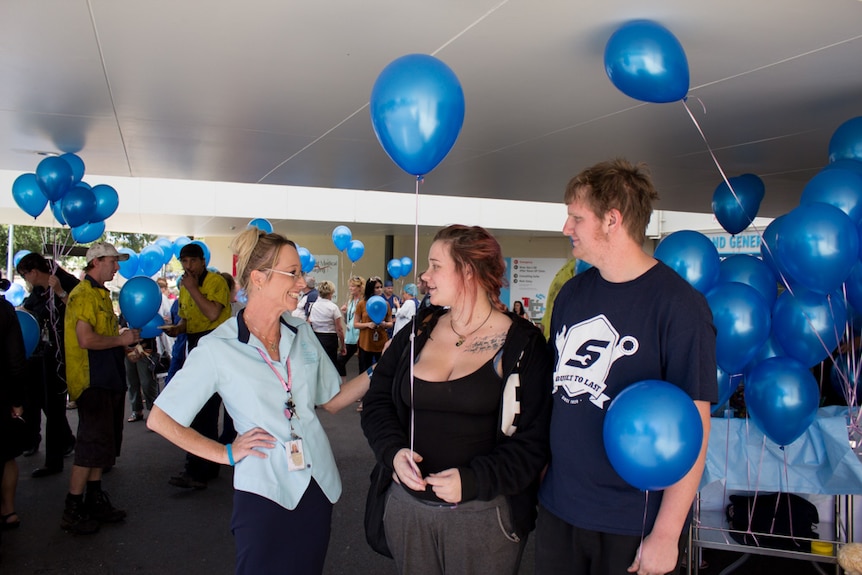 Jennifer Bishop stands with Chloe Sky and Jon Lewis surrounded by people holding blue balloons.