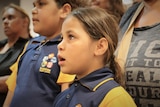 An Indigenous girl in a blue and yellow school uniform singing with a group.